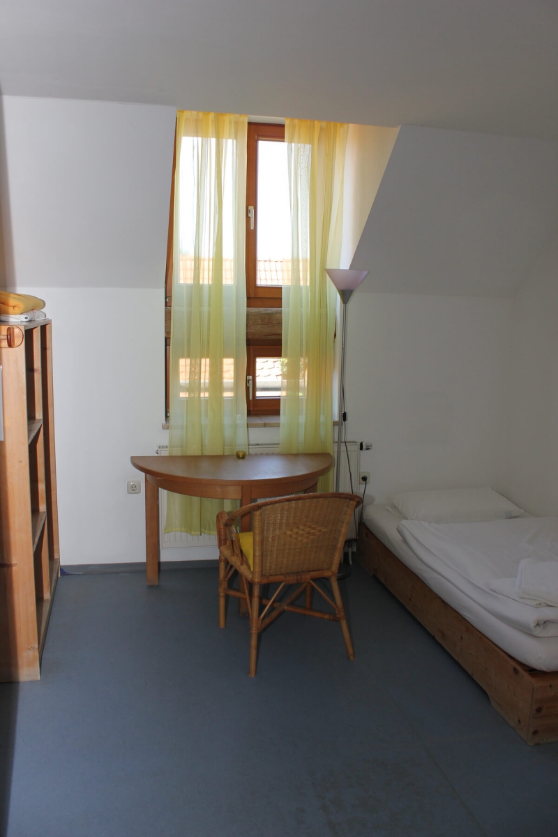 Ein Einzelzimmer mit einem Bett, einer Holzkommode sowie einem kleinen Holztisch inkl. Holzstuhl.